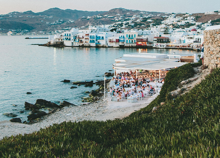 A view of the coast of Mykonos, with a beachside restaurant in the foreground.