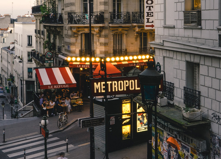 A wide view of a typical city street in Paris, with a busy restaurant and a sign for the Paris metro in view.